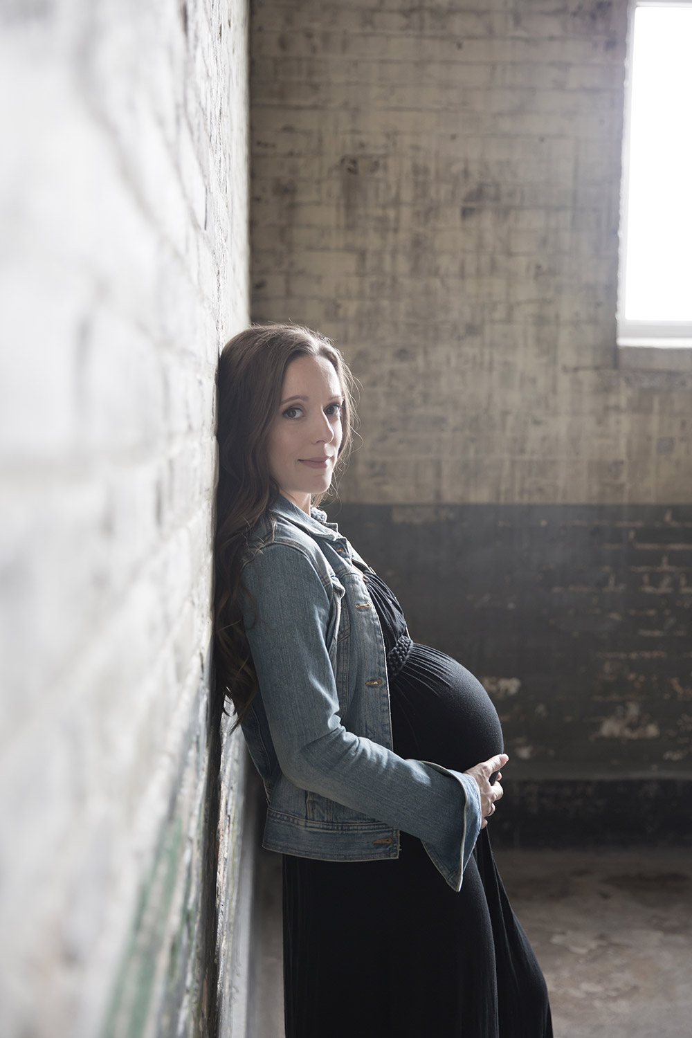 a maternity photo of a woman holding her belly while leaning against an distressed brick wall