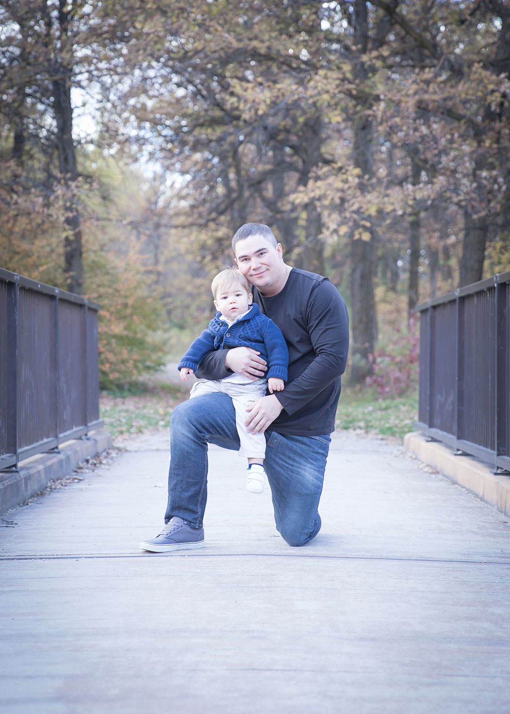 a photo of a little boy sitting on his dad's knee on a bridge