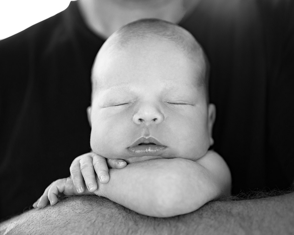 a newborn baby boy posed with his head resting on his arm, while being held by his father