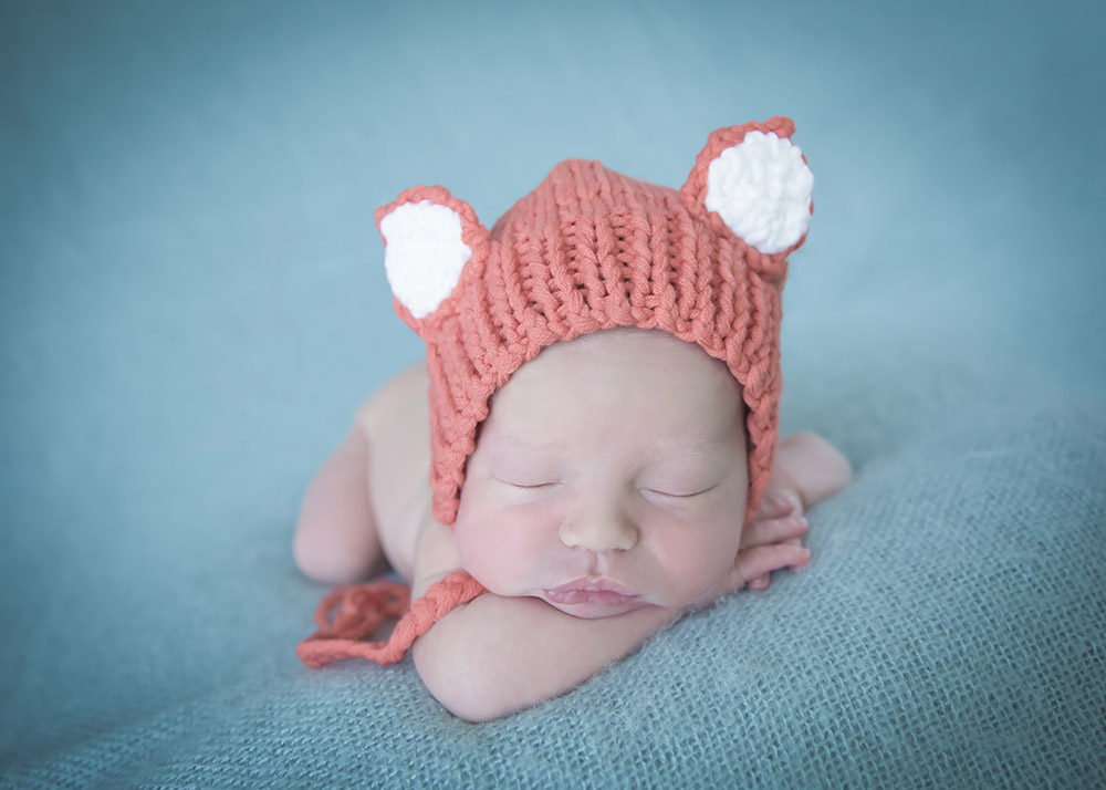a newborn baby boy wearing a fox bonnet, posed on a blue backdrop