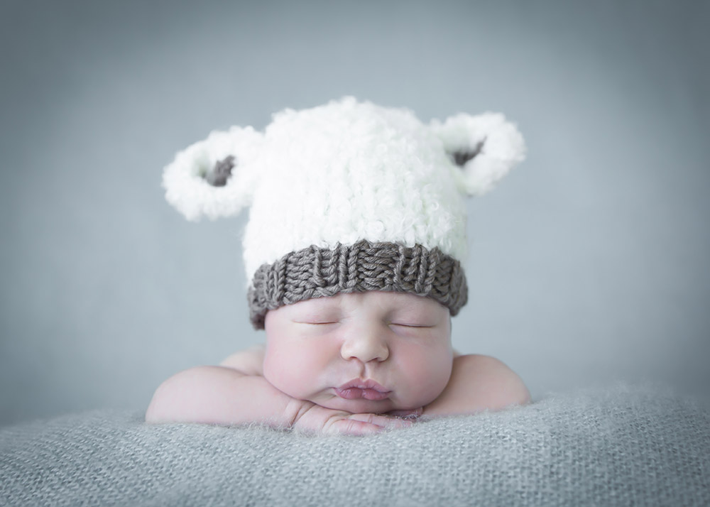 a newborn baby boy, posed with his head resting on his arms, wearing a lamb hat