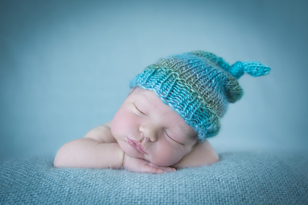 a newborn baby boy posed while sleeping, on a blue backdrop, wearing a knit hat