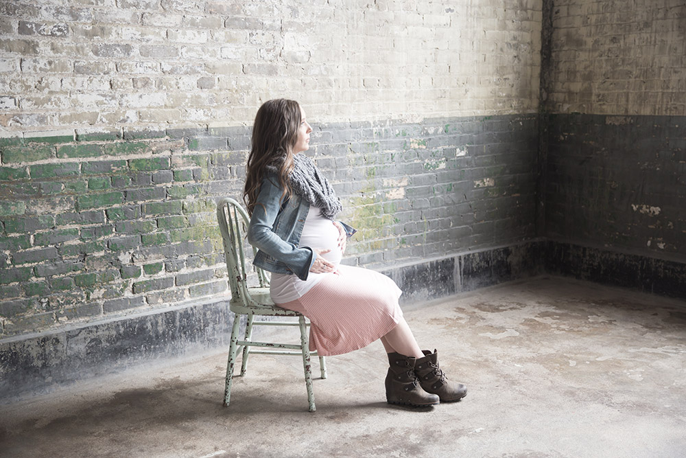 a maternity photo of a woman sitting in a distressed chair looking towards a large window, in front of a distressed brick wall