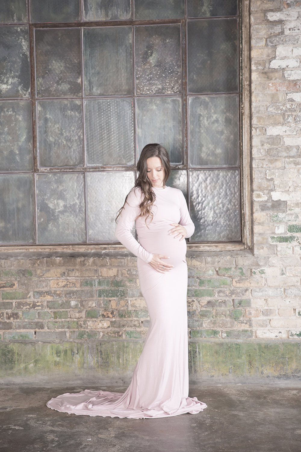 a maternity photo of a woman wearing a long pink gown standing in front of a distressed brick wall and old window