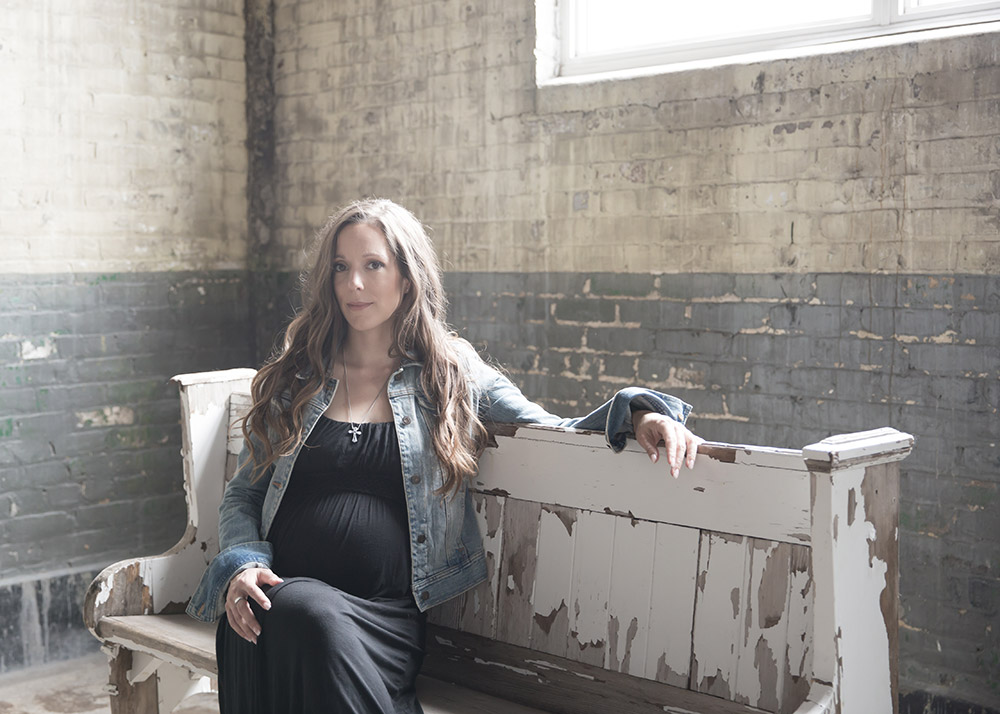 a maternity photo of a woman sitting on an old distressed bench in front of a large window and distressed brick wall