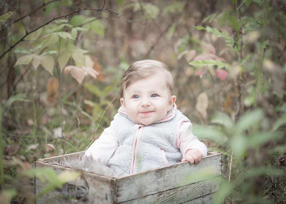 a photo of a baby girl sitting in a wooden crate in the autumn leaves
