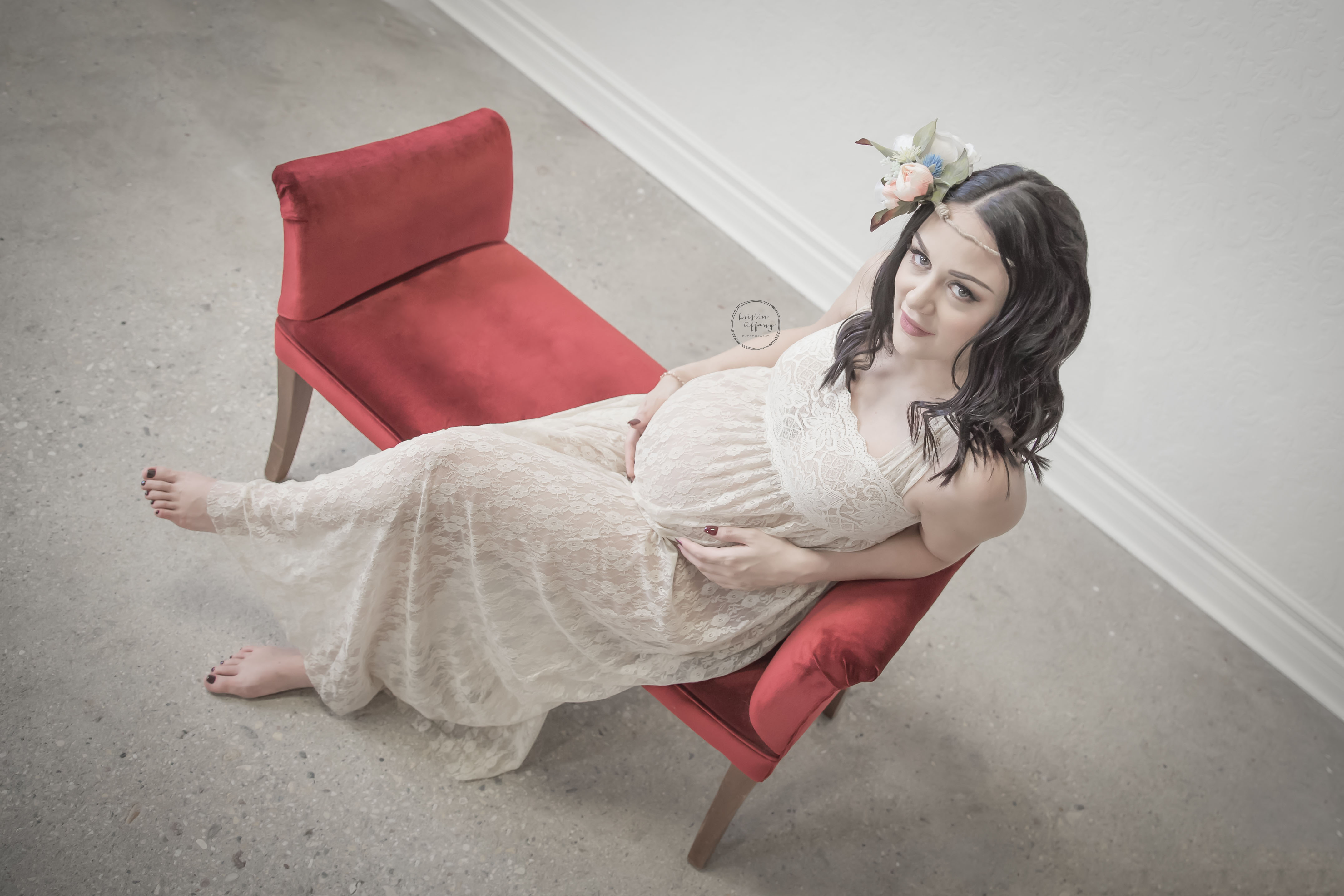 a maternity photo of a woman sitting on a red bench with a lace dress