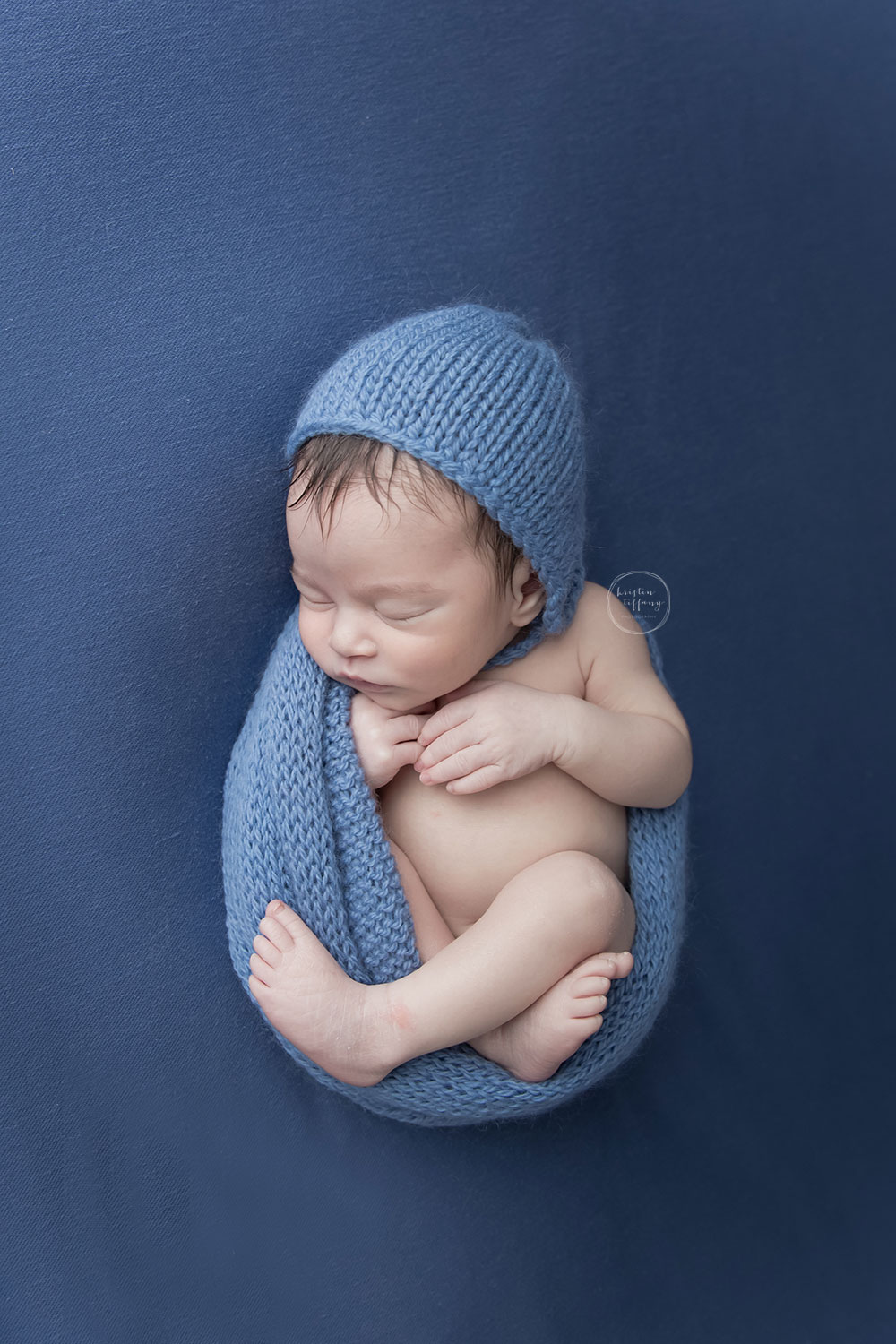 a photo of a posed sleeping baby boy in a blue wrap and bonnet