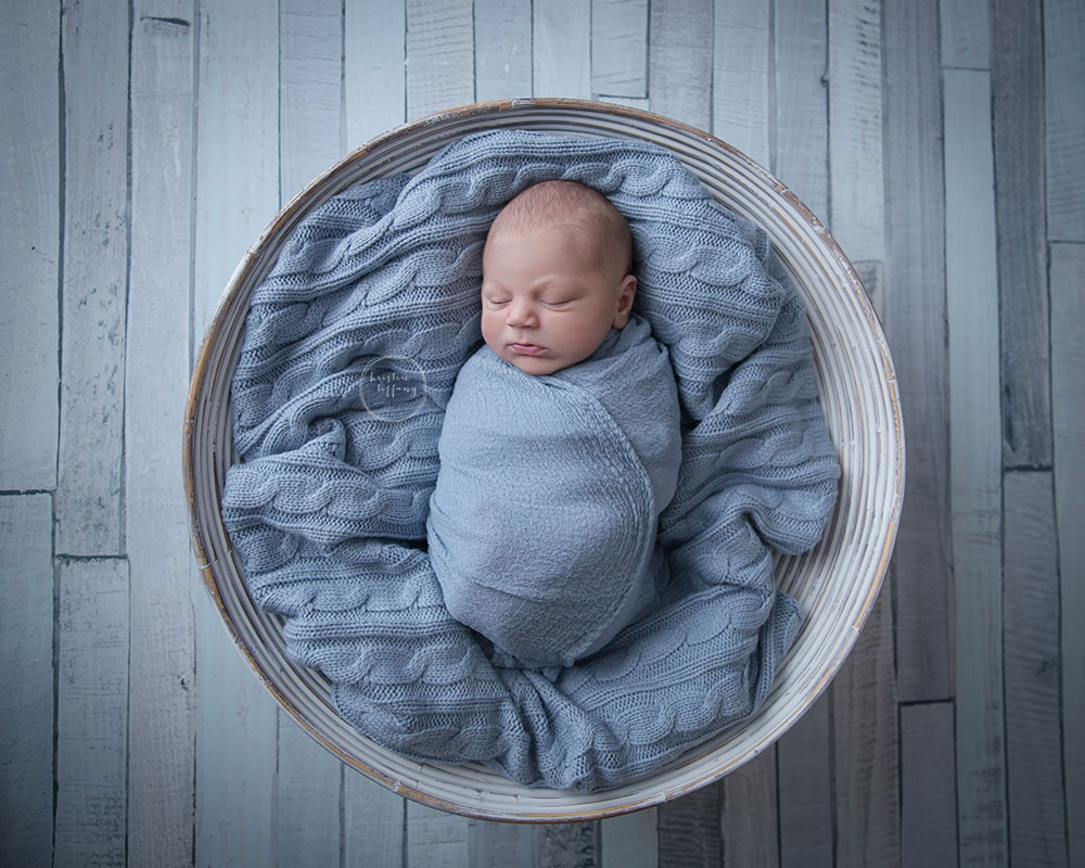 a photo of a newborn baby boy posed in a prop bowl on a wooden floor drop