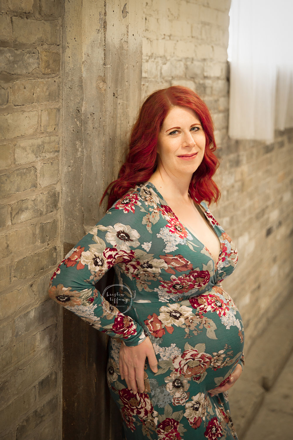 a maternity photo of a woman in a floral dress by a brick wall